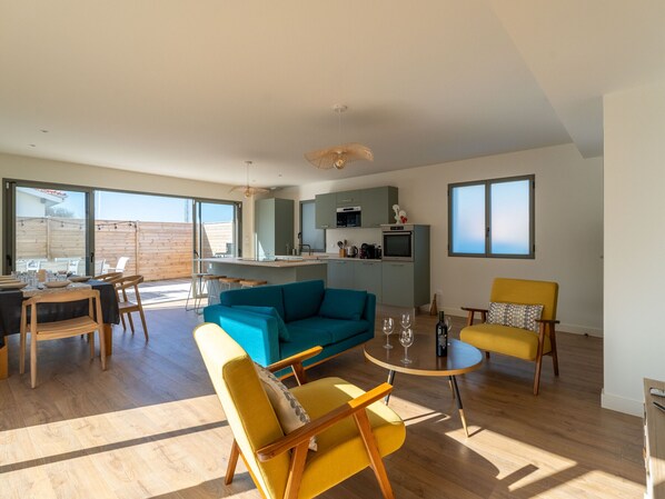 A spacious living area with a sofa, chairs, a dining table, and a kitchen in the background. - New house near ocean, terrace, parking, 8 people (Biscarrosse Plage)