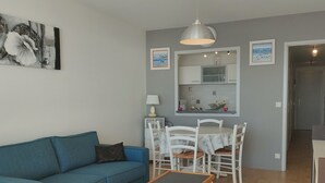 Un salon avec un canapé bleu, une table à manger avec des chaises blanches, une petite table avec une lampe, et un coin cuisine avec un miroir et des placards.