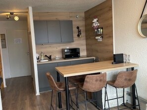 Een compacte keuken met een houten aanrecht, een magnetron en een koffieapparaat. Er zijn twee barstoelen en een klein plankje met keukenspullen.