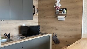Keittiössä on harmaat kaapit, musta mikroaaltouuni, puinen seinä, seinälle ripustettu keittiöpyyhe ja pieni hylly, jossa on kyltti, jossa lukee ”CUISINE”.