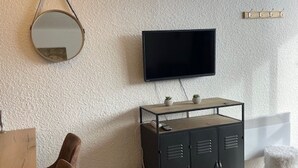 Huoneessa on seinälle asennettu televisio, puinen kaappi, jossa on kaksi pientä kasvia, pyöreä peili ja pehmeä jakkara.