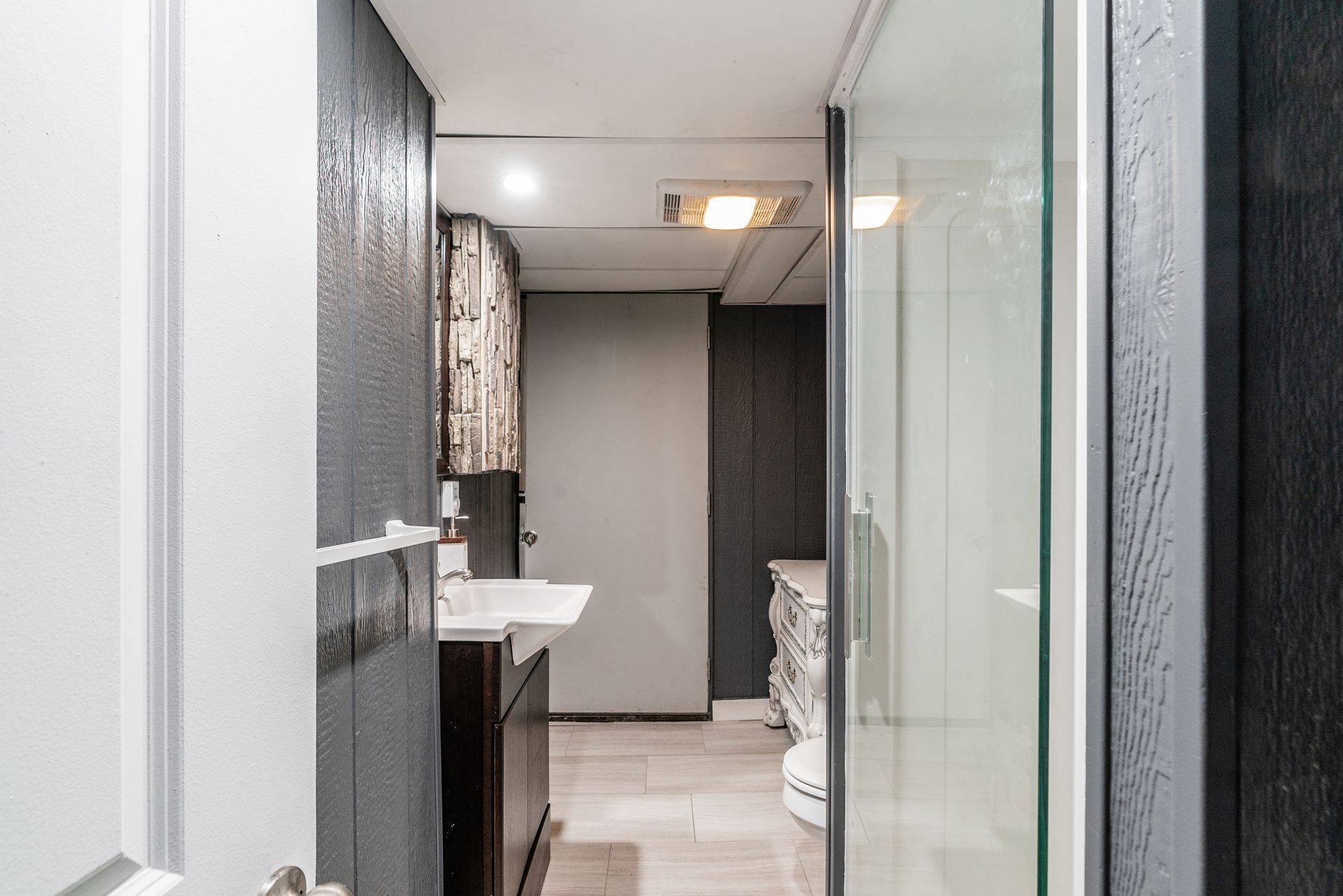 A narrow hallway leading to a bathroom with a white sink, dark wood cabinets, and a glass shower enclosure.