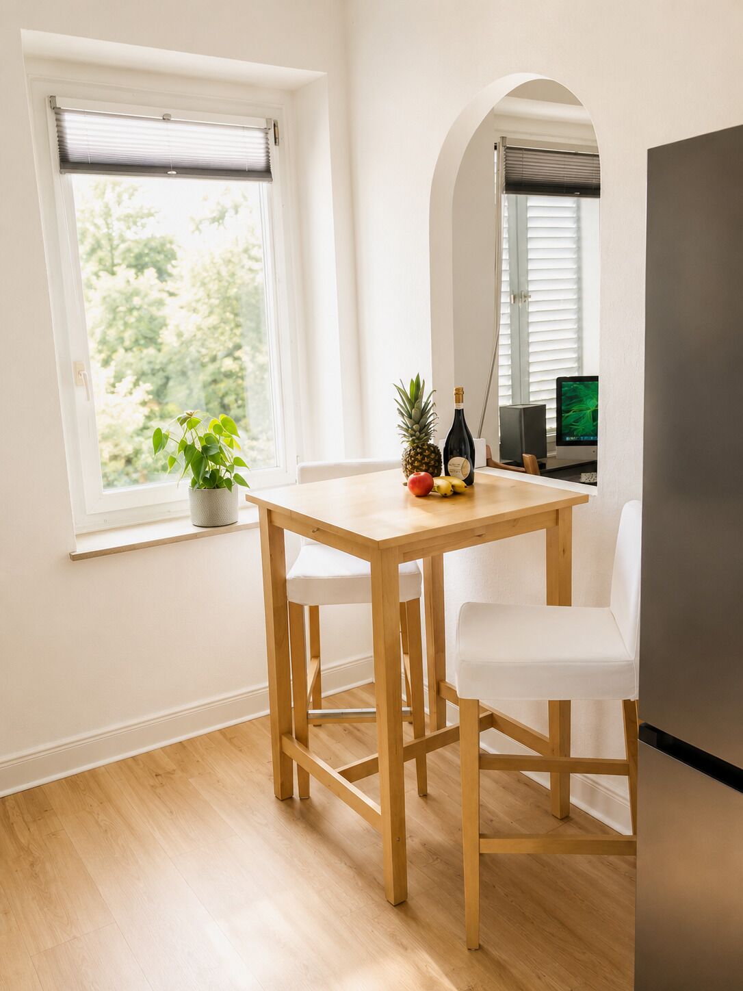 Un petit coin repas avec une table en bois et deux chaises blanches, une plante en pot, une bouteille de champagne et un ordinateur posé sur un bureau.