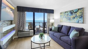 A living room with a sofa, a coffee table, a TV, and a large window with a view of the ocean.