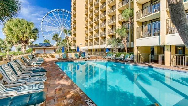A hotel pool area with lounge chairs, a Ferris wheel, and a multi-story building.