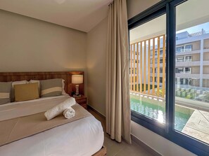 A hotel room with a large bed, a nightstand with a lamp, and a view of a building and a pool through a large window.