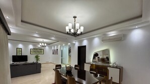 Un comedor moderno con una mesa central, sillas y una araña. Hay una zona de cocina con una encimera y un televisor.