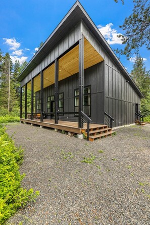 A modern building with a large porch and wooden deck, surrounded by gravel and greenery. - Wagon Wheel Hideaway - All season new build home near Tamarack Resort & Lake Cascade (Donnelly)