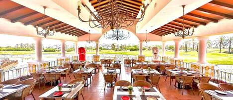 A spacious outdoor restaurant with multiple tables and chairs, a central chandelier, and a view of a landscaped area.