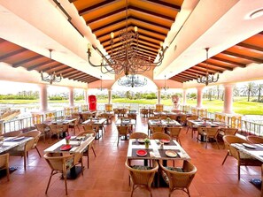 Un vaste restaurant en plein air, doté de nombreuses tables et chaises, d’un lustre central et offrant une vue sur un espace paysager aménagé.