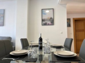 Une table à manger dressée pour quatre personnes, avec des assiettes, des couverts et des verres à vin, une bouteille de vin et un tableau encadré accroché au mur derrière.
