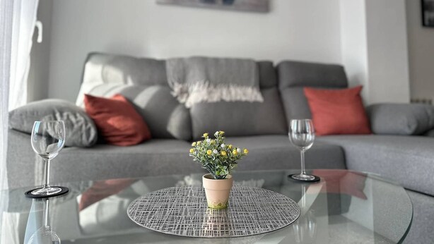 A living room with a glass coffee table, two wine glasses, a potted plant, and a sofa with cushions. - Bright & modern 2 bedroom apartment - TR721LT (La Torre Golf Resort)