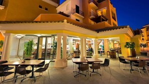 An outdoor dining area with tables and chairs, a covered section with a glass wall, and a building with multiple balconies and a staircase. - Bright & modern 2 bedroom apartment - TR721LT (La Torre Golf Resort)