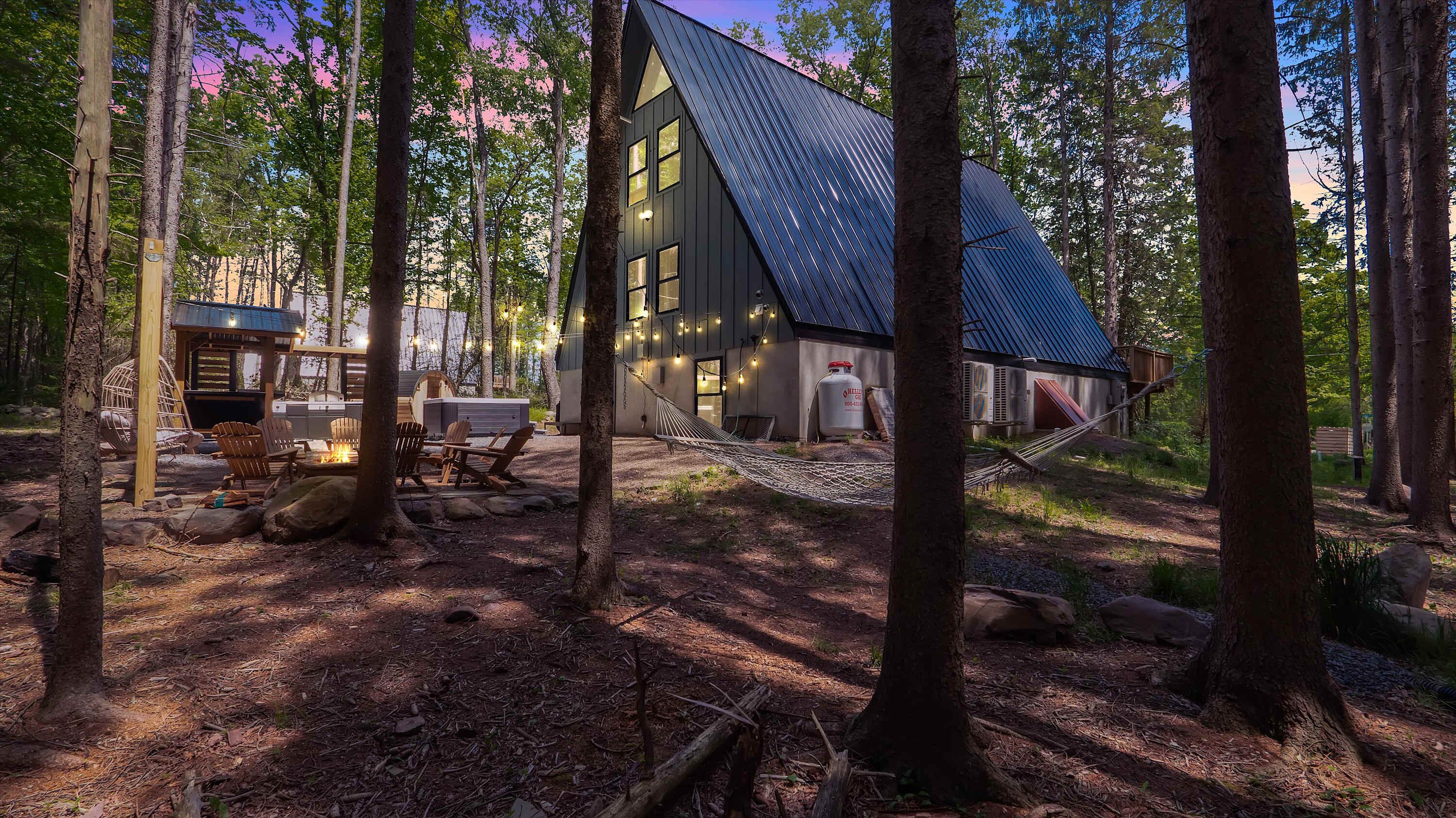 A cabin with a sloped roof, surrounded by trees, featuring a fire pit and outdoor seating.