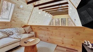 Ein gemütliches Blockhaussinnenzimmer mit Holzbalkendecke, einer Couch, einem runden Tisch und Blick durch ein Fenster.
