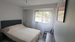 A bedroom with a bed, a nightstand, a chair, and a window with sheer curtains.