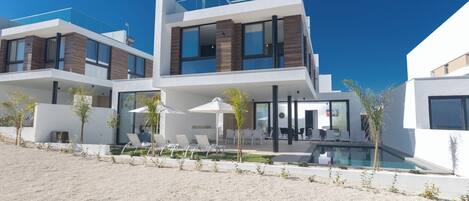 Una casa moderna de dos pisos con piscina, mobiliario exterior y un cielo azul despejado.