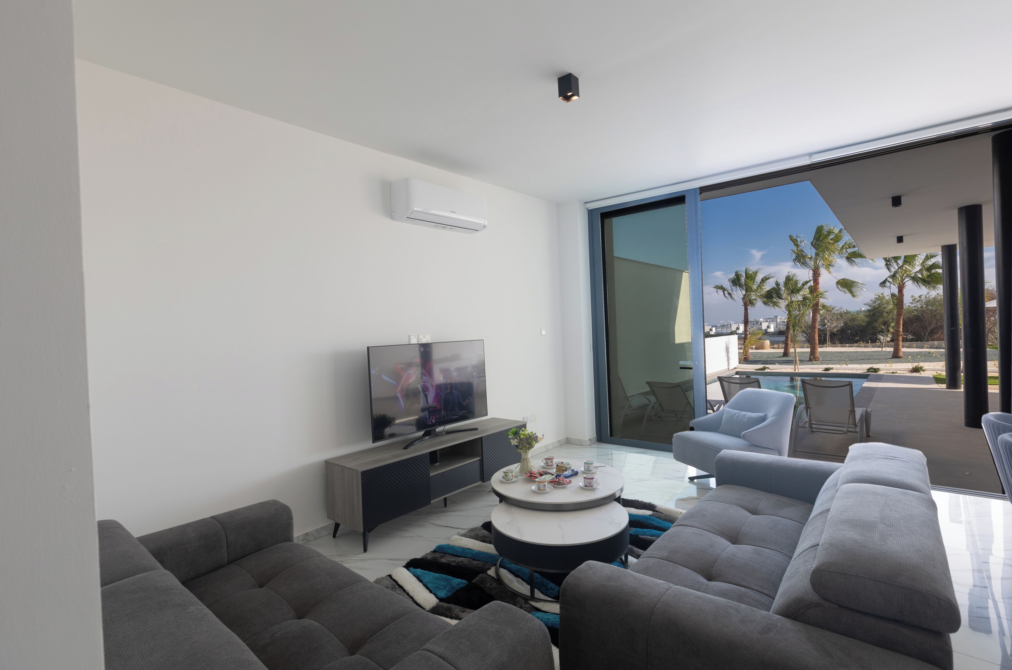 Una sala de estar moderna con una televisión de pantalla plana, sofás grises, una mesa de centro y un ventanal de vidrio que da a un balcón con piscina y palmeras.