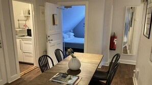 Un comedor con una mesa de madera, cuatro sillas y un jarrón con flores. Se ve una cama al fondo.