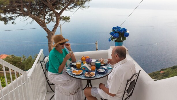 Una pareja cenando en una mesa con vista al mar, una planta en maceta y una barandilla blanca.