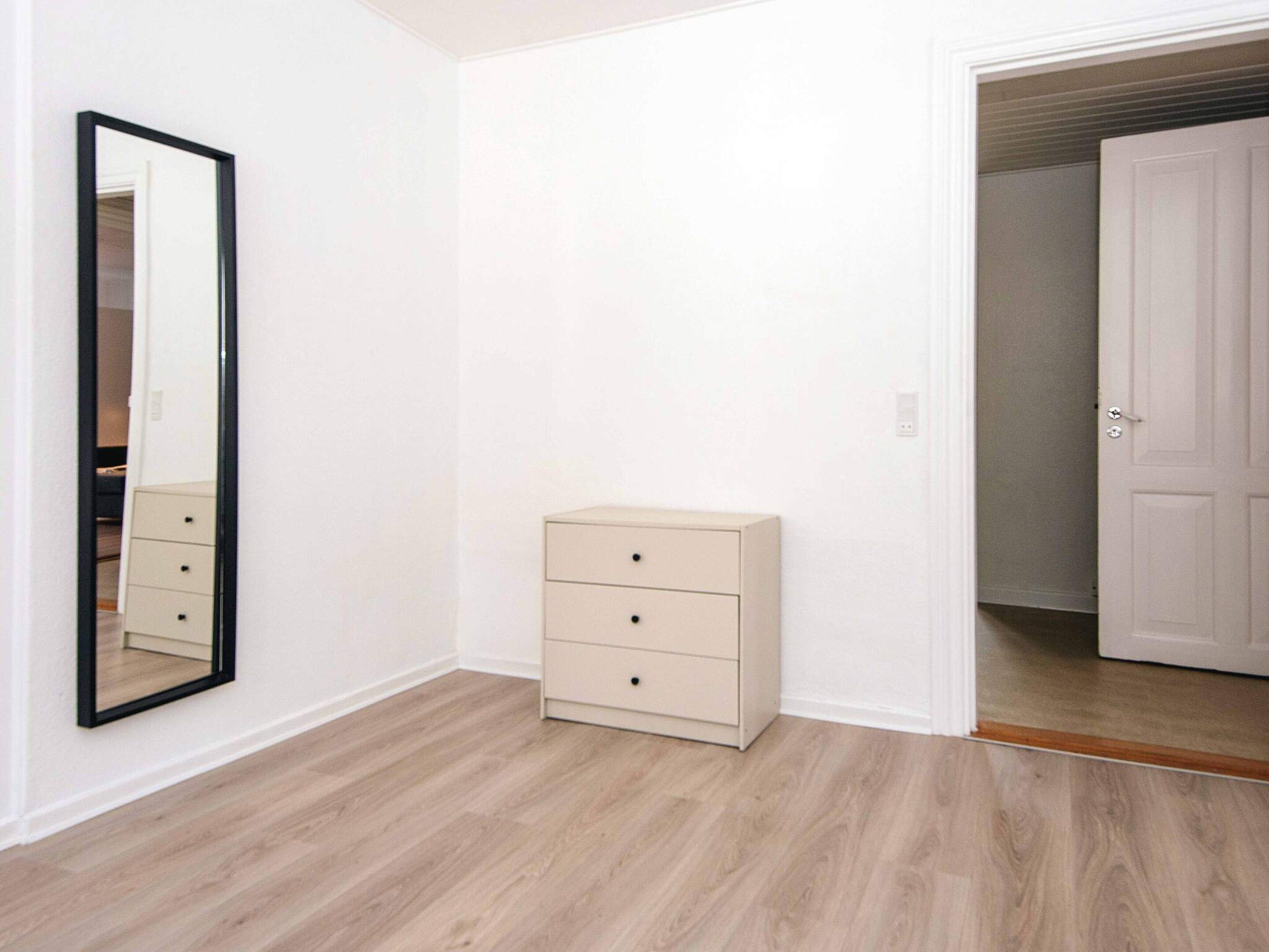 Una habitación con piso de madera, un aparador beige con cajones, un espejo de cuerpo entero y una puerta blanca que da a otra habitación.