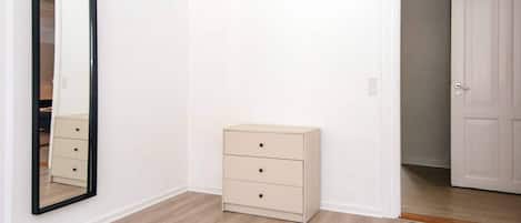 Una habitación con piso de madera, un aparador beige con cajones, un espejo de cuerpo entero y una puerta blanca que da a otra habitación.