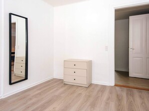 A room with a wooden floor, a beige dresser with drawers, a full-length mirror, and a white door leading to another room.