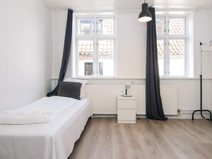 Une chambre à coucher avec un lit, une table de chevet, une lampe, un radiateur et une fenêtre avec des rideaux.