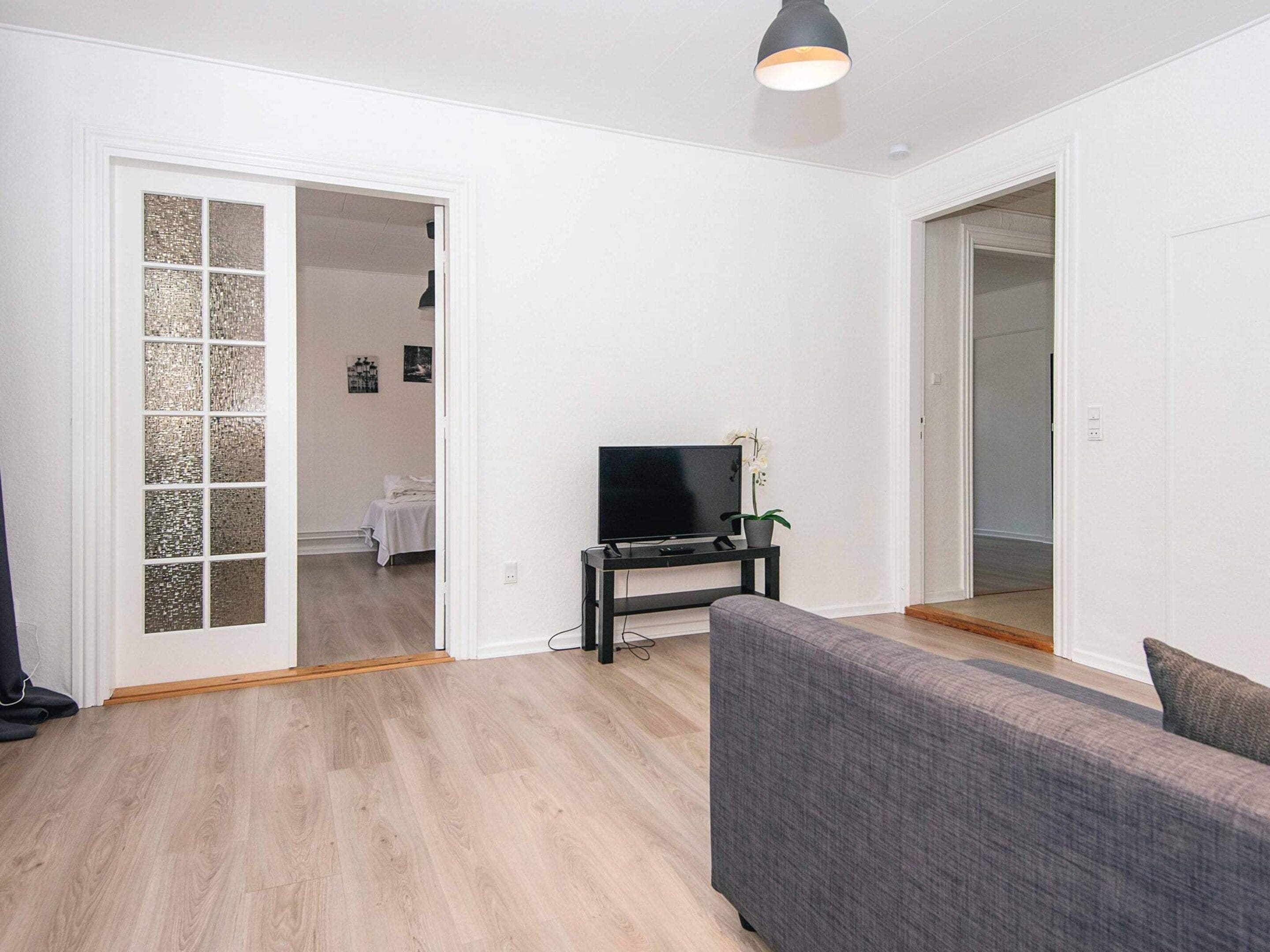 Un salon avec un canapé gris, un téléviseur posé sur un meuble et une porte donnant sur une autre pièce.