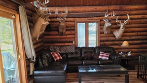 L’intérieur d’une cabane en rondins, avec un plancher en bois, un canapé en cuir, une table basse en verre et deux têtes de cerf fixées au mur.