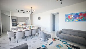 A modern living room with a grey sofa, a dining area with a white table and grey chairs, a kitchen with grey cabinets, and a colorful abstract painting on the wall.