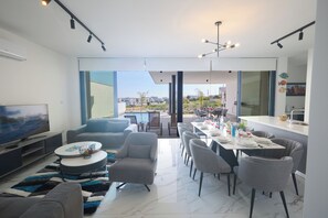 A modern living room with a TV, seating, and a dining area with a view. - Protaras Aqua Pearl Villa Aq12 (Paralimni)