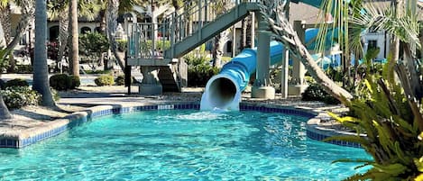 A swimming pool with a blue slide, surrounded by palm trees and a building with a balcony.