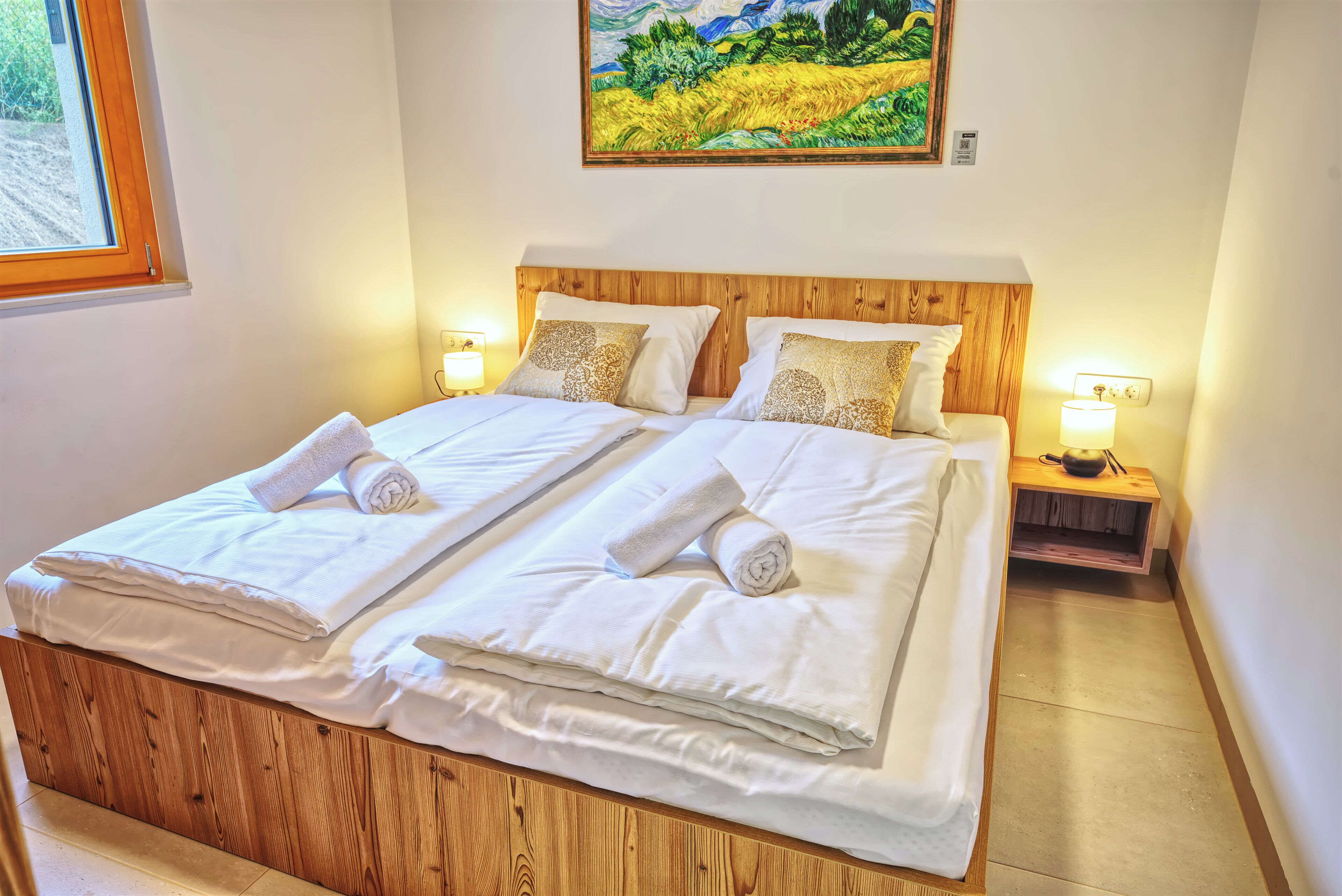 Una cama doble con sábanas blancas y dos almohadas, un cabecero de madera, mesitas de noche con lámparas y un cuadro sobre la cama.