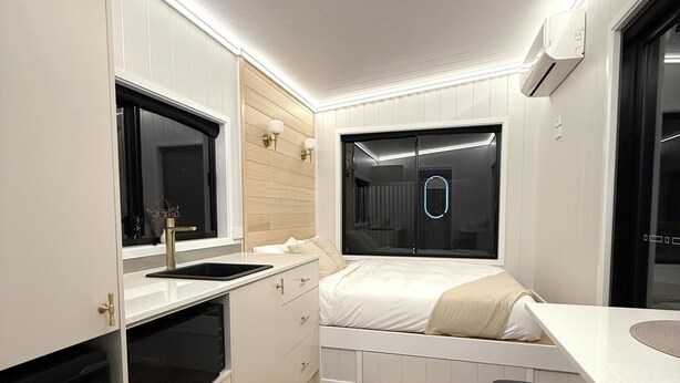 A compact, modern kitchenette with built-in appliances, a small dining area, and a bed with a window view.