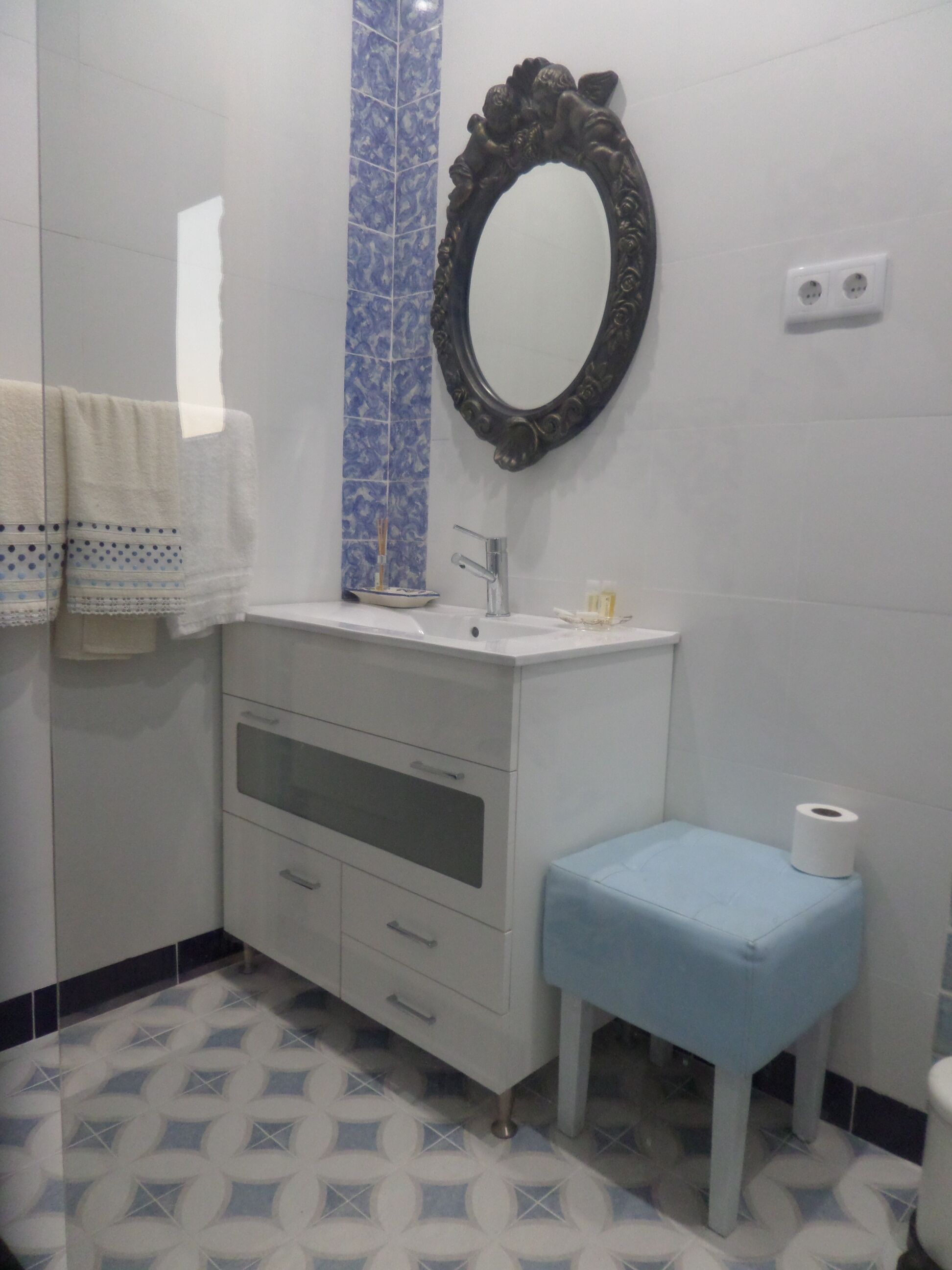 A bathroom with a white vanity, a blue-tiled column, a mirror, a toilet, and patterned floor tiles.
