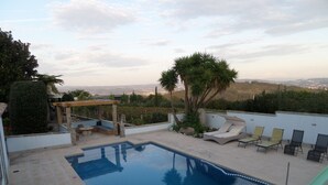 Ein Swimmingpool mit Liegestühlen und einer Pergola, umgeben von Grünflächen und Blick auf ferne Hügel.
