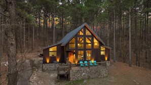 A cabin with a stone foundation, large windows, and a deck with chairs. - NEW! Prime Cabin Chateau, Hot Tub-Firepit-Sleeps 6 (Broken Bow)