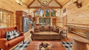 A log cabin interior with a wooden ceiling, a chandelier, a mounted deer head, a brown leather sofa, and a dining area. - NEW! Prime Cabin Chateau, Hot Tub-Firepit-Sleeps 6 (Broken Bow)