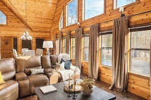 A log cabin interior with a leather sofa, a chandelier, and large windows with curtains. - As Seen on TV! Safari Views + Hot Tub + Firepit (Broken Bow)