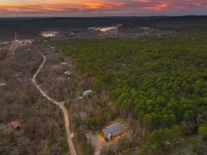 An aerial view of a forested area with a dirt road, several buildings, and a small lit structure. - As Seen on TV! Safari Views + Hot Tub + Firepit (Broken Bow)