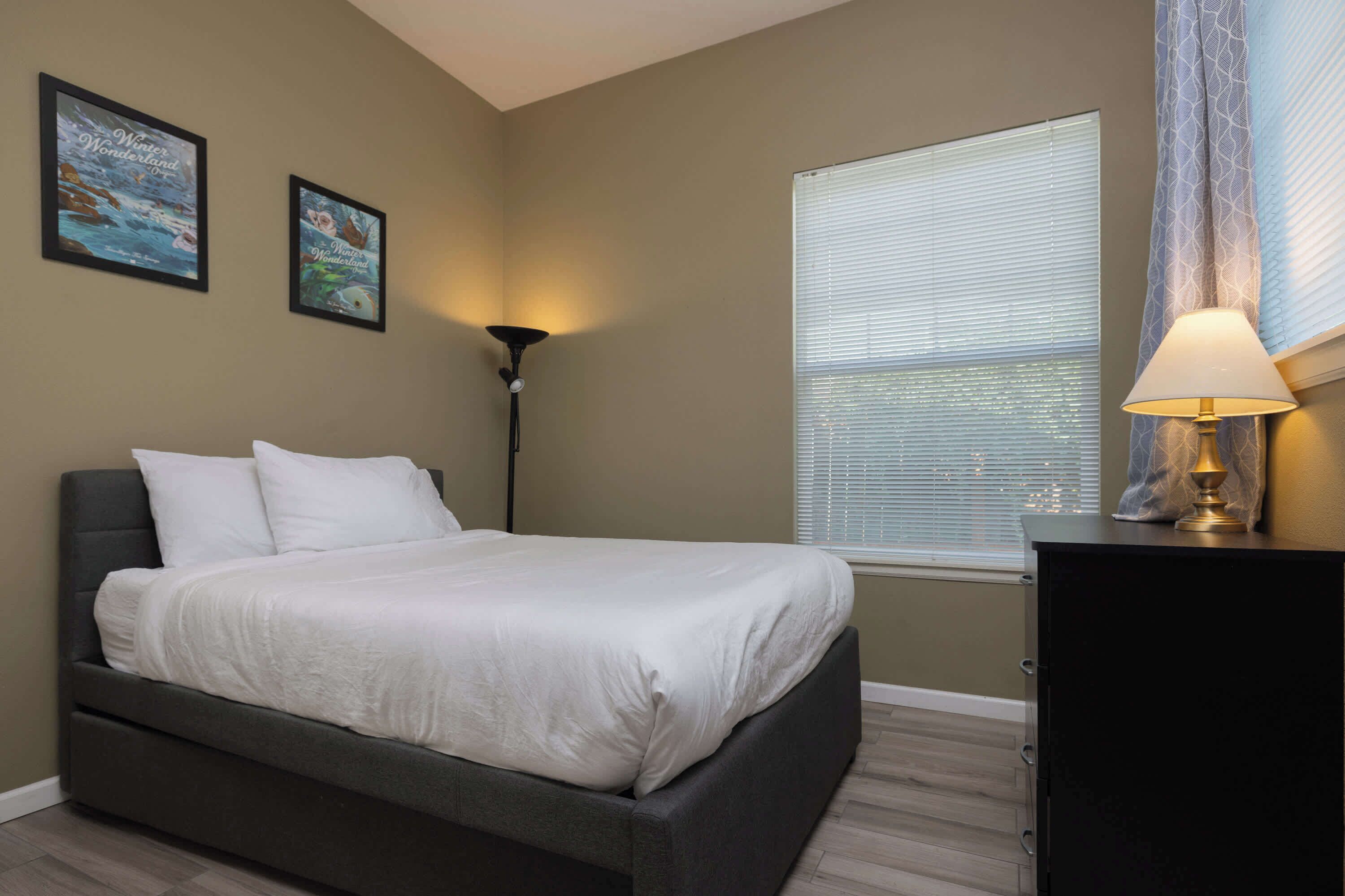 A bedroom with a bed, a lamp, a window with blinds, and two framed pictures on the wall.