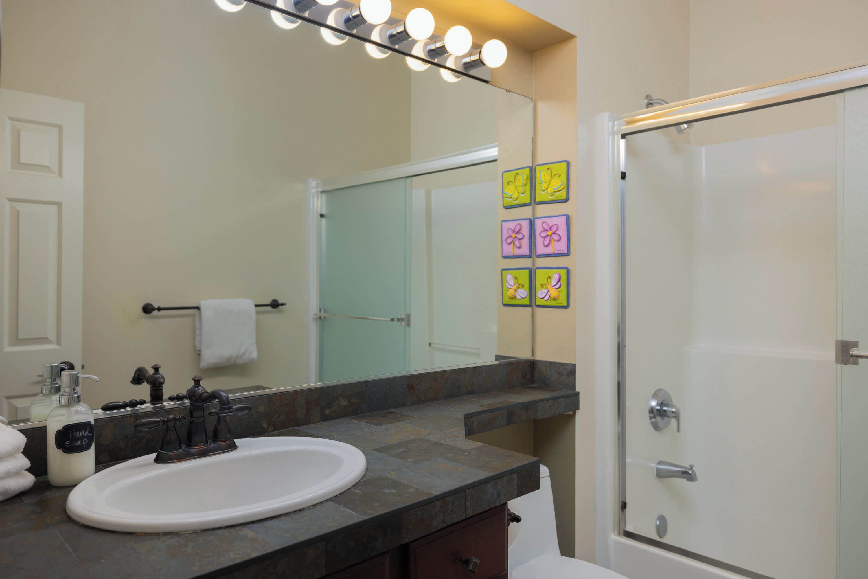 A bathroom with a white sink, a mirror with lights, a glass shower door, and a towel rack.