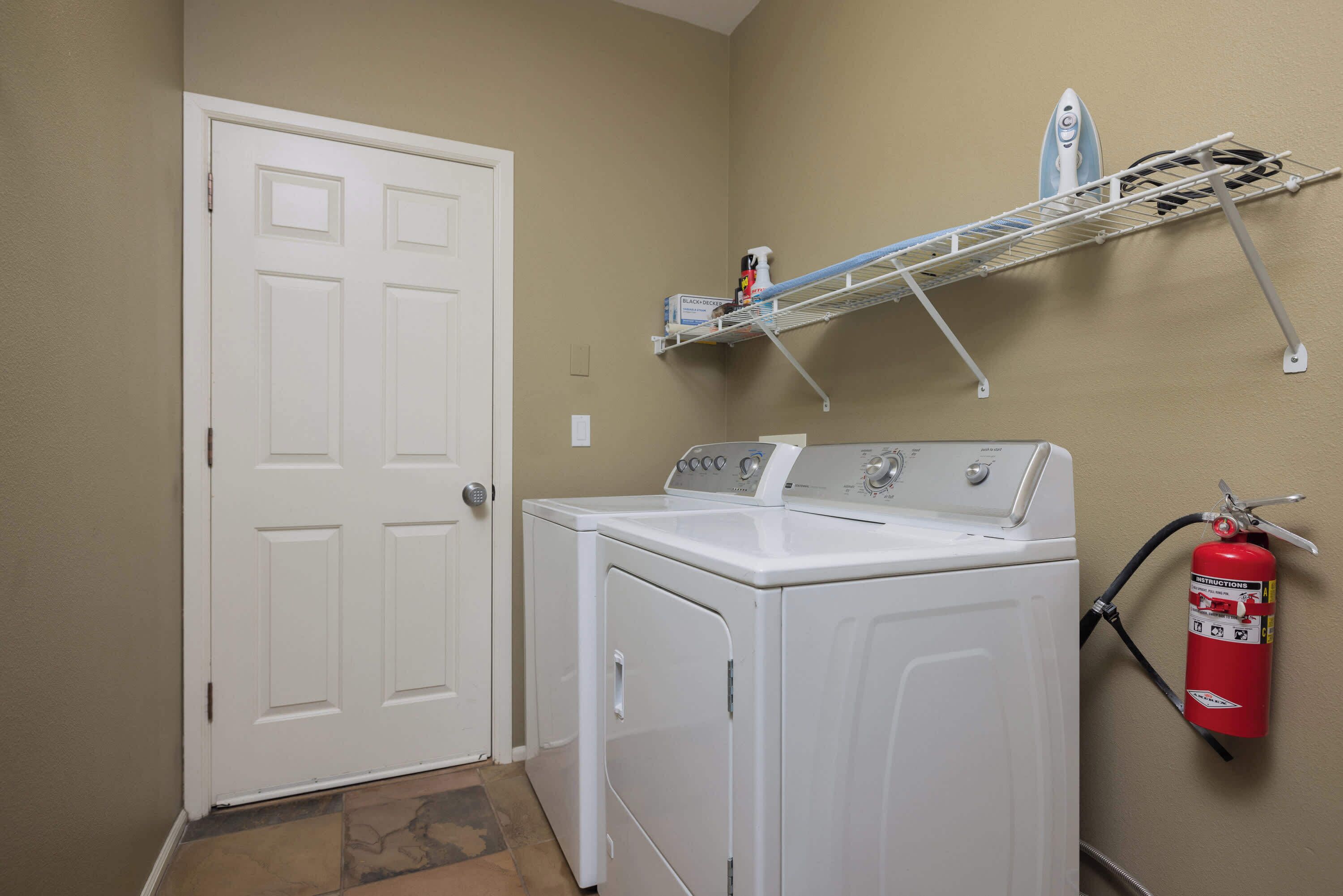 A laundry room with a white door, a washing machine, a dryer, a shelf with cleaning supplies, and a fire extinguisher.