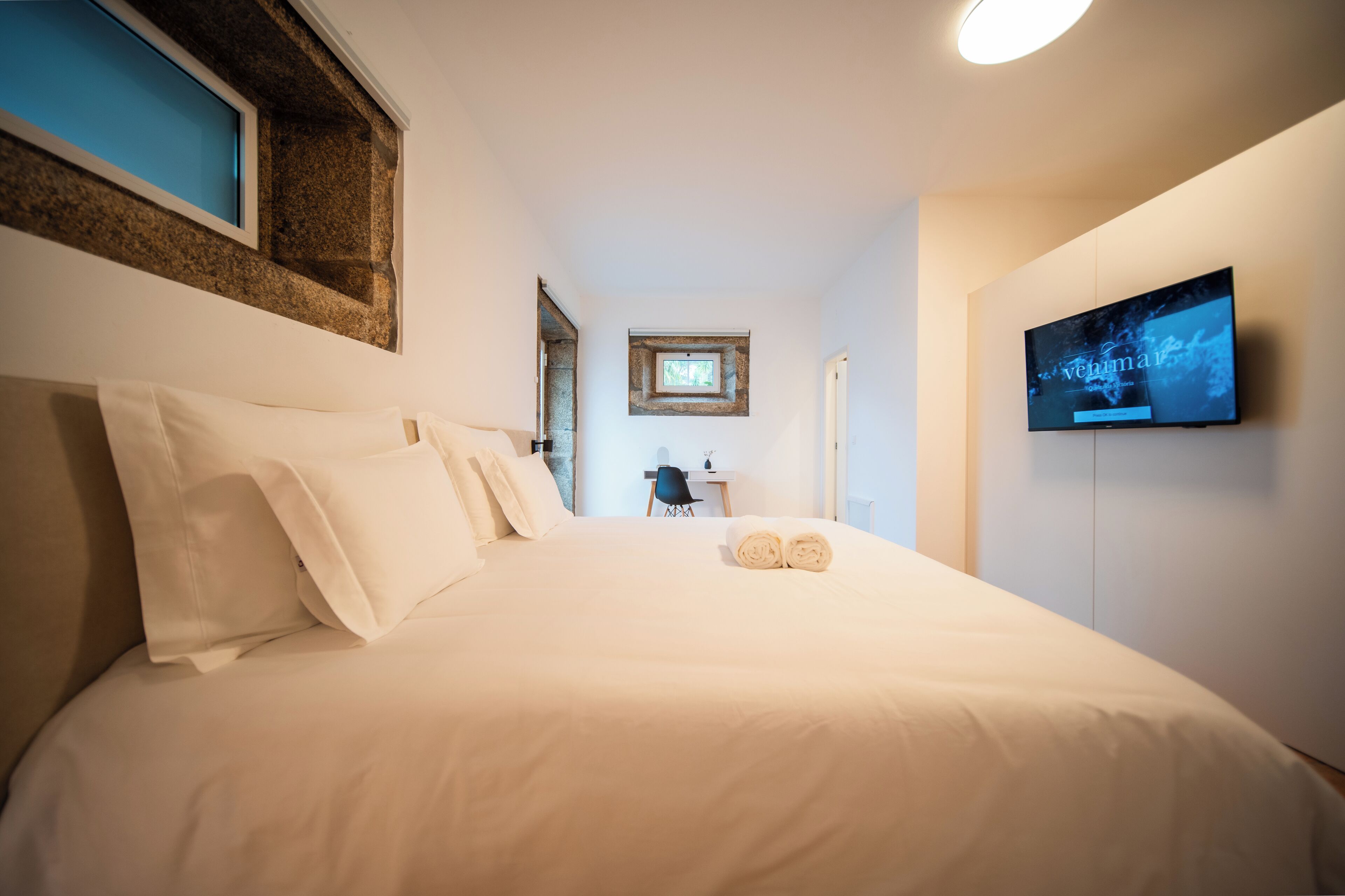 A hotel room with a large bed, a desk, a chair, a TV, and a small window.