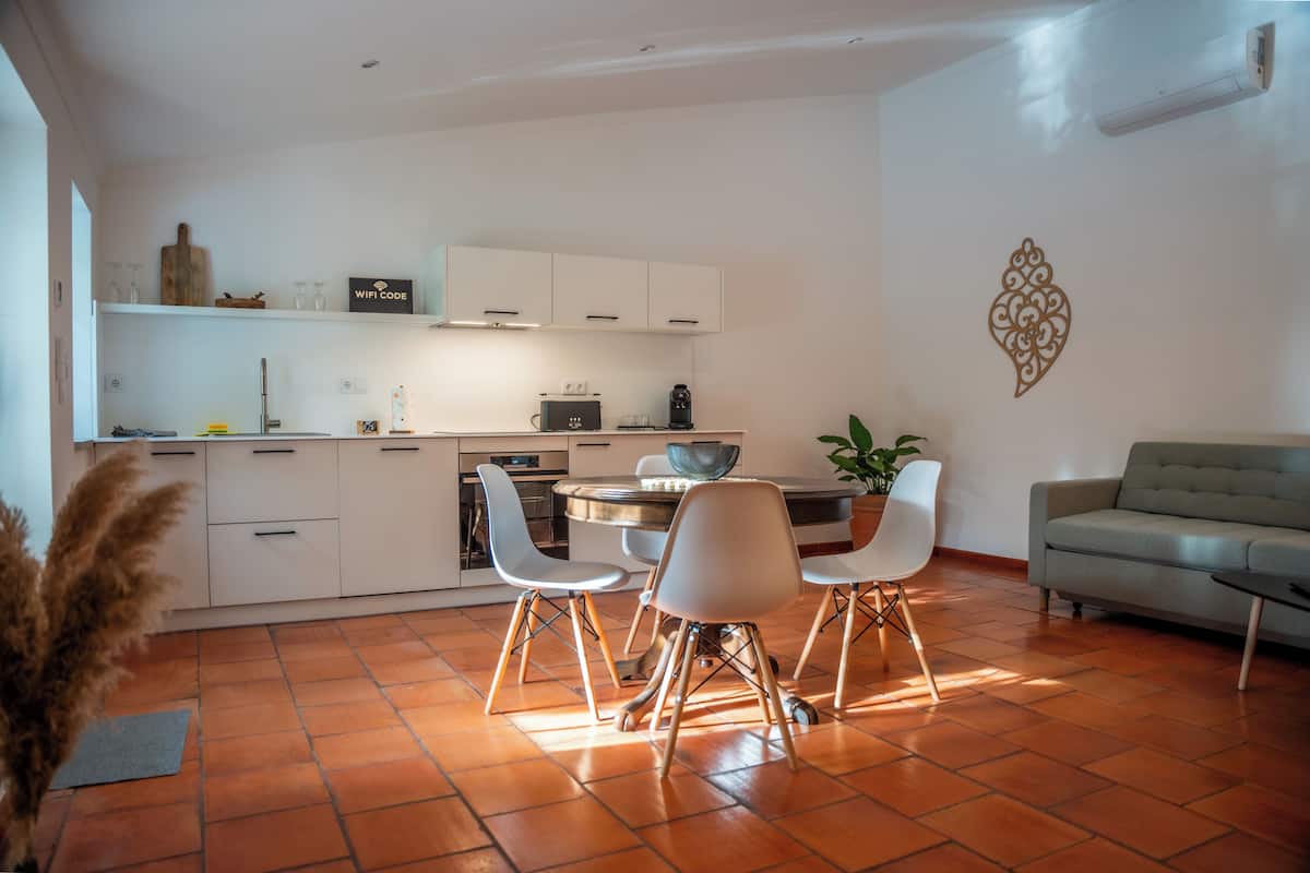 A modern kitchen with white cabinets, a dining area with white chairs, and a grey sofa.