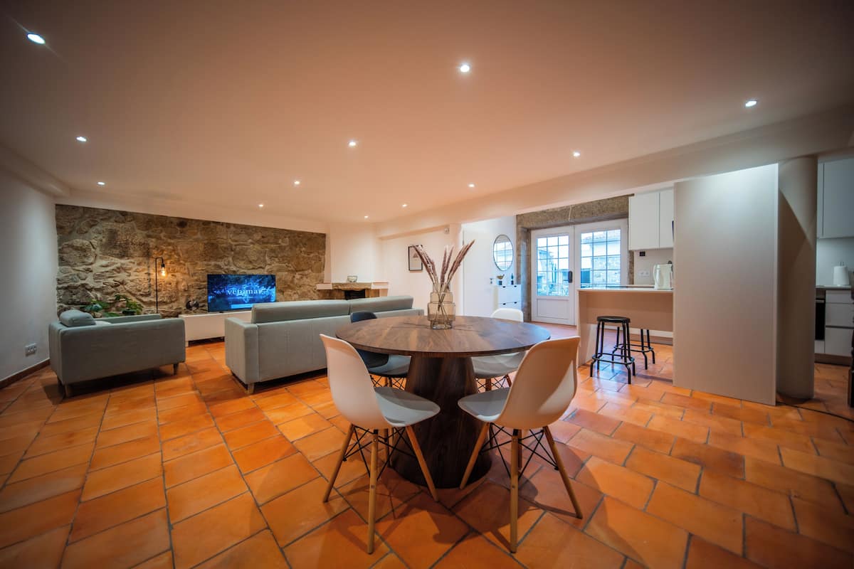 A modern living room with a stone wall, a round dining table with chairs, a grey sofa, and a TV.