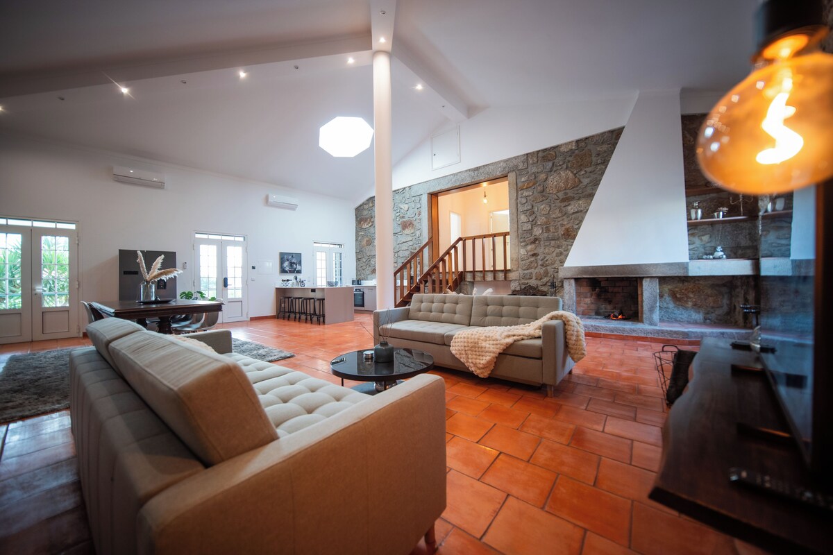 A spacious living area with a sofa, coffee table, and a fireplace. There is a staircase leading to another level, and a kitchen area is visible in the background.
