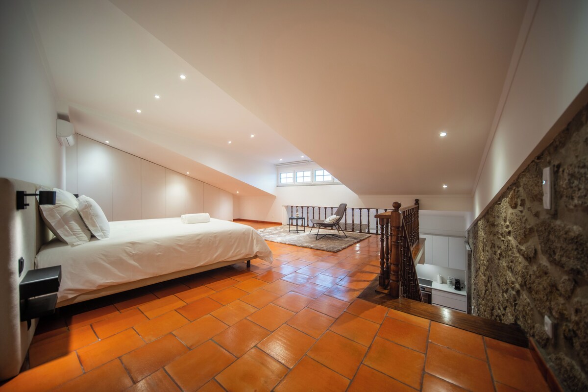A bedroom with a bed, a wooden staircase, a stone wall, and a view of the kitchen.