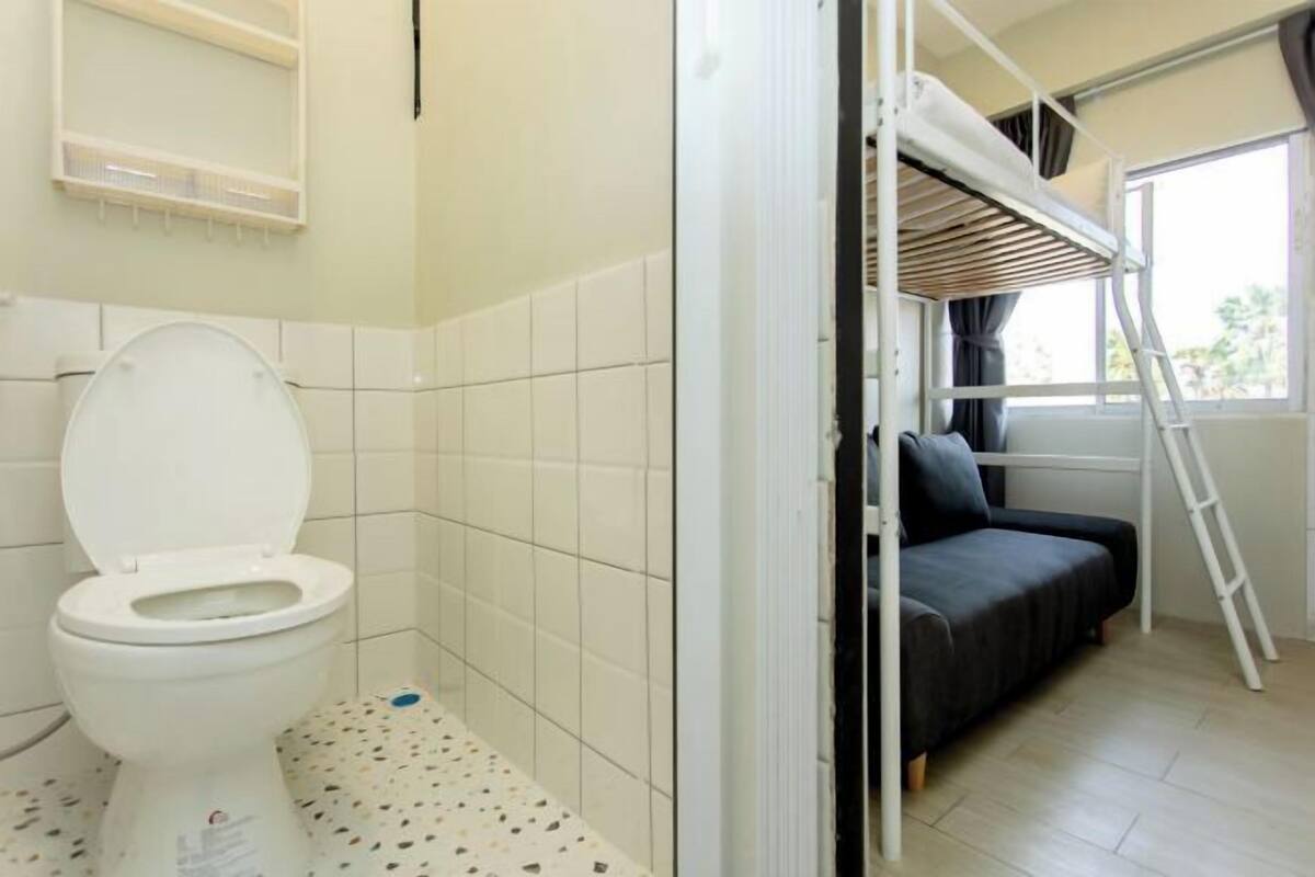 A compact room with a toilet, a bed with a ladder, and a window.